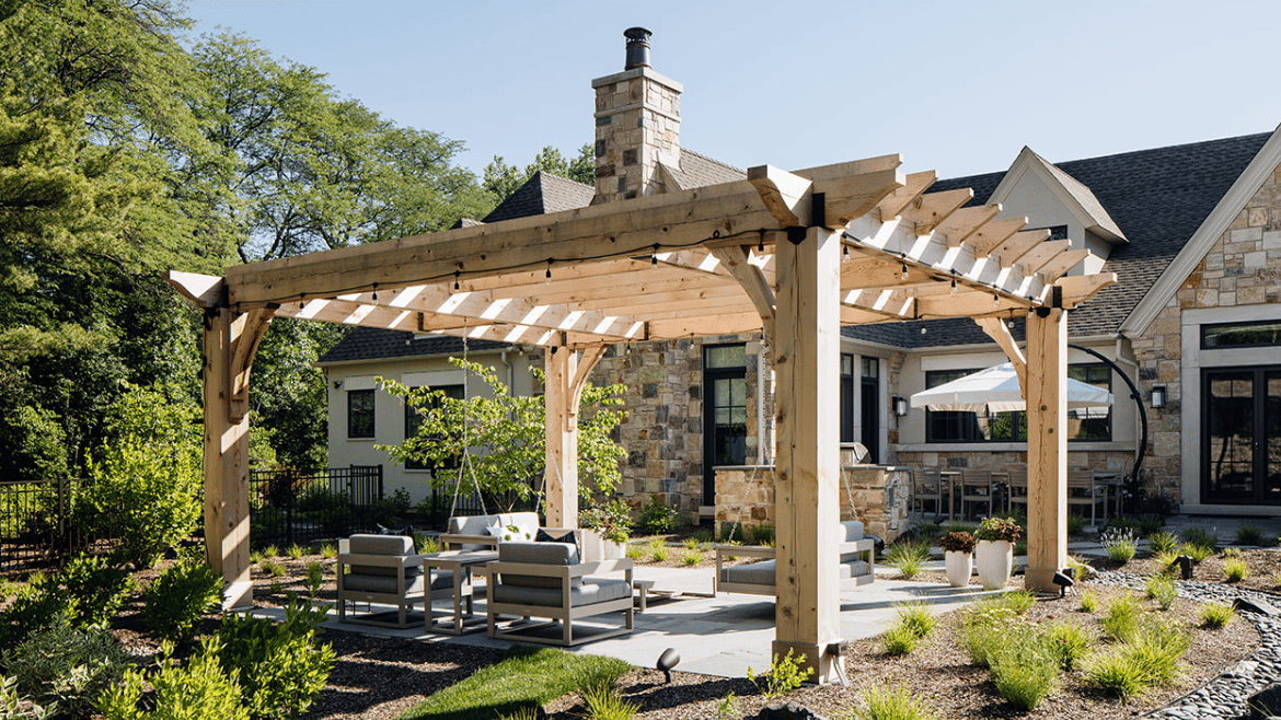 Wood pergola seating area of French Country Style Home with couch swings, blue sky, stone walkway and plantings