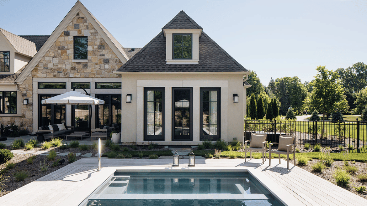 Pool deck of a French Country Style Home with blue sky over two roof peaks, stone facade, dark trim on doors, stone walkway