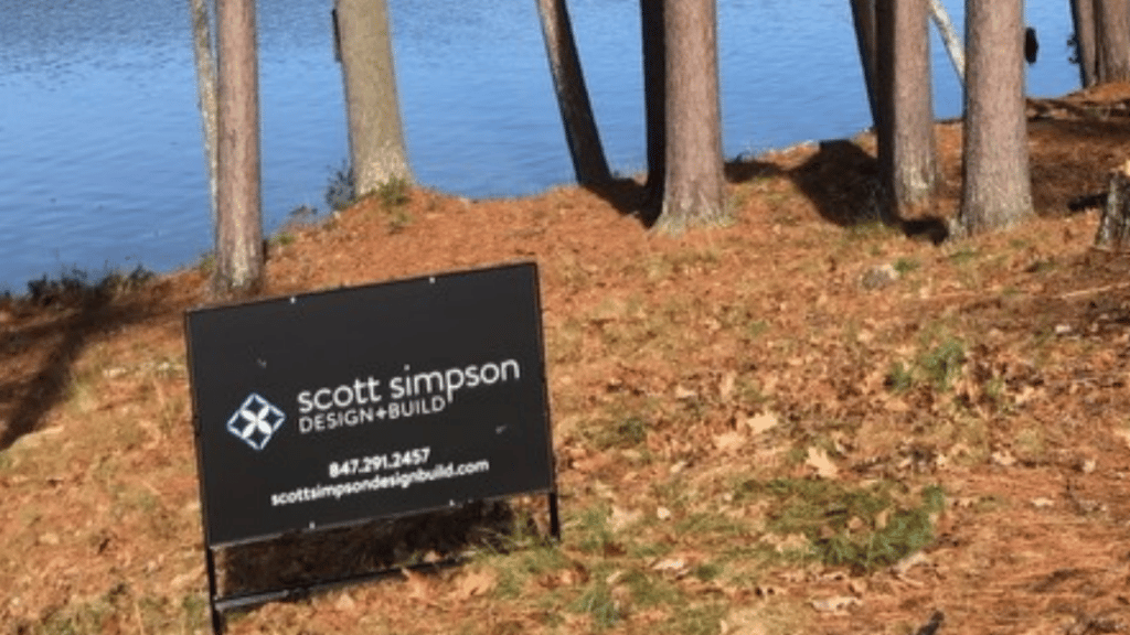 Scott Simpson Design Build metal sign in fall leaves by a lake new home construction build site