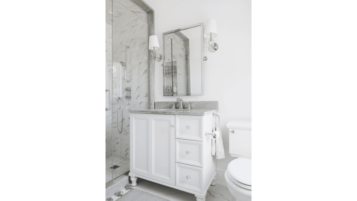 Farmhouse white and gray bathroom vanity with crystal pulls and white lampshade sconces next to gray marble and glass shower